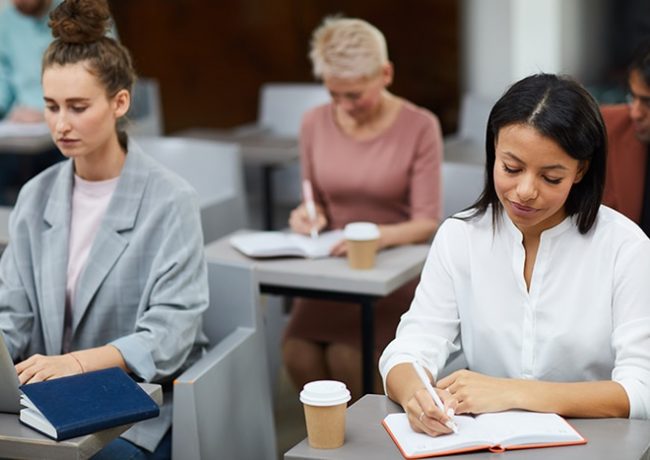 Portrait of mixed-race businesswoman writing notes while sitting at table in class with group of students, copy space
